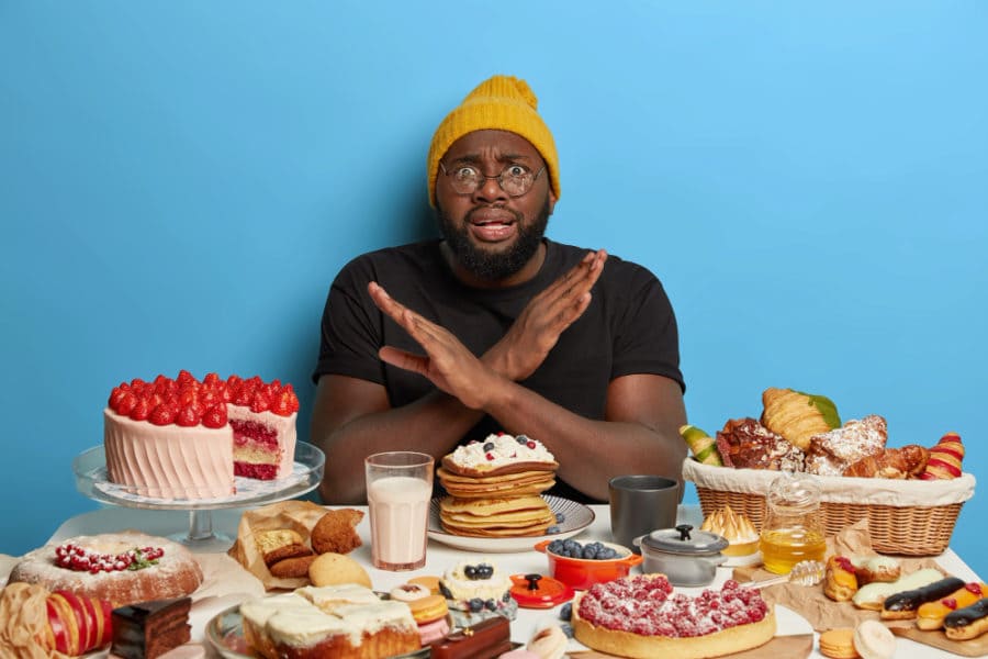 African american man crosses arms over chest, makes denial gesture, refuses eat sugary products, sits at table with bakery, keeps to diet 